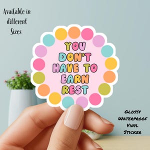 May include: A glossy, waterproof vinyl sticker with the text "YOU DON'T HAVE TO EARN REST" in colorful bubble letters. The sticker has a pink background and is surrounded by a scalloped edge with colorful polka dots. The words "Available in different Sizes" are also visible.
