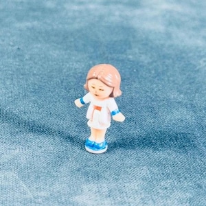 May include: A miniature figurine of a girl with short brown hair, wearing a white dress with blue accents on the sleeves and shoes. The figurine is standing on a blue surface.