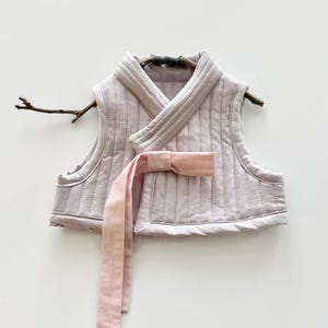 May include: A light purple quilted vest with a wrap-around design and a matching pink sash. The vest is displayed on a small branch against a white background. The vest has a V-neck and a bow closure.
