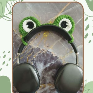 Könnte beinhalten: Grüne gehäkelte Frosch-Kopfhörerabdeckungen mit weißen Augen. Die Abdeckungen sind an einem schwarzen Kopfhörer befestigt.  "CROCHET PATTERN BY AEROL KNOX" steht unter den Kopfhörern.  PDF WRITTEN PATTERN steht in der oberen rechten Ecke.