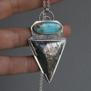 May include: A silver pendant necklace featuring a turquoise stone and a triangular, metallic stone. The turquoise stone is oval-shaped and set above the triangular stone. The pendant hangs from a delicate silver chain.