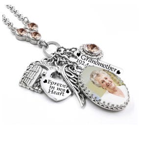 May include: A silver-toned charm necklace featuring a photo pendant of an elderly person, a heart charm engraved with "Forever in my Heart", a garden gate charm, an angel wing charm, and a rectangular tag reading "Grandmother 1924".