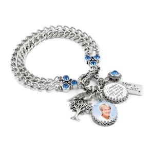 May include: A silver-toned double chain bracelet featuring blue crystal accents, a tree charm, a wing charm, and two circular pendants. One pendant displays a photograph of a person, while the other bears the text: Sometimes I just look up smile, and say, I know that was you. A rectangular tag reads: Mom 1946-2017.