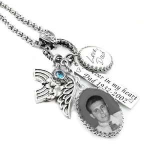 May include: A silver-toned memorial necklace featuring a chain, a blue crystal-accented angel wing charm, a rectangular bar engraved with "Forever in my heart Dad 1932-2002", and two oval pendants, one with a black and white photo and one with "Love, Dad".