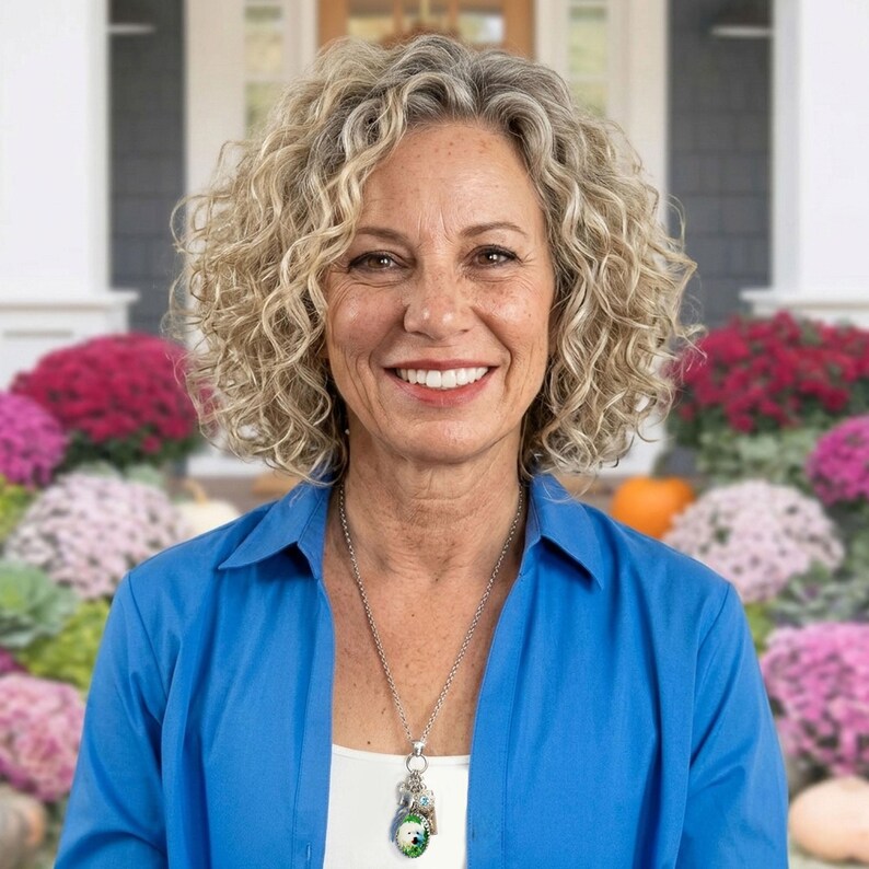 A smiling woman with curly blonde hair wearing a blue blazer and a necklace stands in front of a house with colorful flowers in the background.