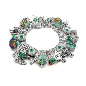 May include: A silver-toned charm bracelet featuring multiple circular charms with colorful illustrations, green faceted beads, and rectangular metal tags engraved with the text Dorothy, Glinda the Good Witch, and OZ, along with a red shoe charm.