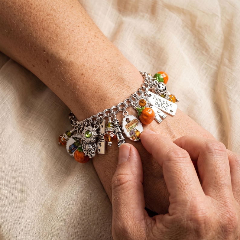 May include: A silver-tone charm bracelet featuring various autumn-themed pendants including orange pumpkins, green and amber beads, a scarecrow, and a rectangular tag engraved with the text Pumpkin Patch and another with Fall Harvest.