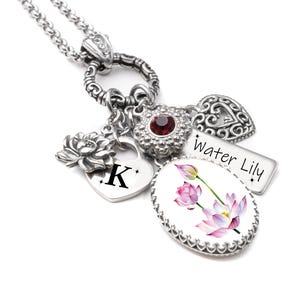 May include: A silver-toned necklace featuring a central ring with multiple charms: a heart charm with the letter K, a silver lotus flower, a flower charm with a red center stone, a heart-shaped filigree charm, and a rectangular tag labeled Water Lily.