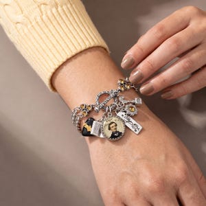 May include: A silver-toned charm bracelet featuring multiple dangling elements, including a circular pendant with a portrait of Edgar Allan Poe, a rectangular tag inscribed with his name, a black heart, a book, and small amber-colored crystals.