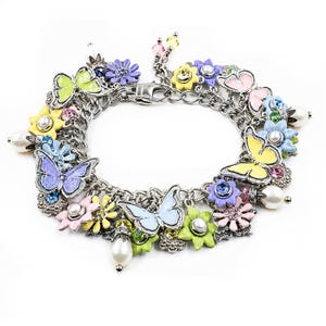 Op de afbeelding: Een zilverkleurige bedelarmband met een verscheidenheid aan kleurrijke emaille vlinders en bloemen. De bedels zijn geaccentueerd met kleine witte kunstparels, heldere kristallen en levendige tinten geel, roze, blauw, groen en paars emaille.