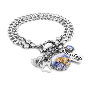 May include: A silver-toned chain link bracelet featuring a toggle clasp, clear rhinestones, a silver paw print charm, a small dog figurine, a blue crystal, and a circular pendant with a golden dog image and a rectangular tag reading Bailey 2001-2017.
