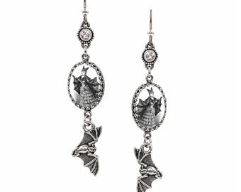 May include: A pair of silver-toned drop earrings featuring a circular clear crystal stud, an oval pendant displaying a black and white illustration of a figure in a bat-winged costume, and a dangling silver bat charm at the base of each earring.