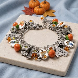 May include: A silver-tone charm bracelet featuring various autumn-themed charms including orange pumpkins, green and amber crystals, and rectangular metal tags with text such as Pumpkin Patch, Scarecrow, and Falling Leaves.