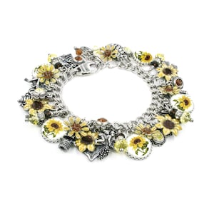 May include: A silver-toned charm bracelet featuring multiple sunflower-themed charms, including yellow and brown enamel sunflower pendants, round sunflower illustrations, and small amber-colored crystal beads attached to a thick chain.