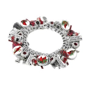 Op de afbeelding: Een zilveren bedelarmband met meerdere ronde hangers met illustraties van rode kardinaalvogels, rode kristallen kralen en rechthoekige metalen plaatjes gegraveerd met de teksten Your wings already exist, Angels watching over you en I am always with you.
