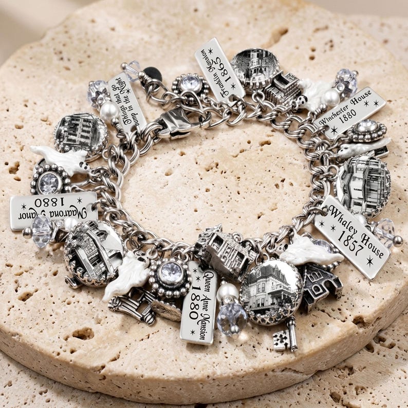 May include: A silver charm bracelet featuring multiple dangling charms, including black and white circular images of historic houses, white ghost figures, clear faceted beads, and rectangular metal tags inscribed with names and dates like Franklin Mansion 1865, Winchester House 1880, Whaley House 1857, Queen Anne Mansion 1880, and Things that go bump in the Night.