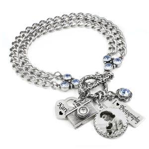 May include: A silver-toned double chain bracelet featuring light blue crystal accents, a toggle clasp, and multiple charms including a camera, a heart with the name Kari, a rectangular tag with the text I heart Photography, and a circular photo charm.