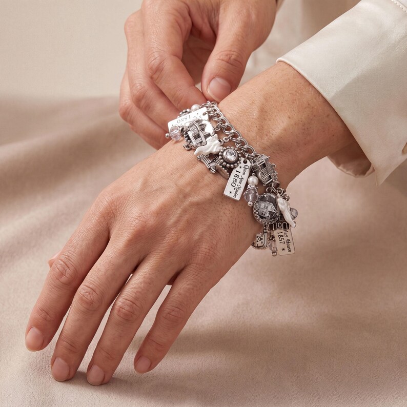 May include: A silver-toned charm bracelet featuring multiple dangling ornaments, including small house-shaped charms, clear faceted beads, and rectangular metal tags engraved with the text Queen Anne Mansion 1880 and Bailey House 1857.