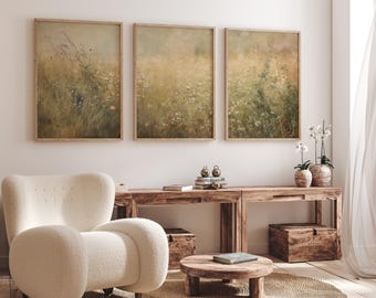 May include: Three framed landscape art prints with a vintage aesthetic. The artwork depicts a field of wildflowers in muted tones of green, yellow, and brown. The prints are displayed above a wooden console table, with a cream-colored armchair in the foreground.