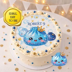 Peut inclure: Un gâteau rond avec une décoration comestible représentant un personnage de boulette bleue portant une couronne dorée, le texte "ROBERT 9" et des bulles bleues. Le gâteau est orné de tourbillons de glaçage blanc, de perles et de paillettes.