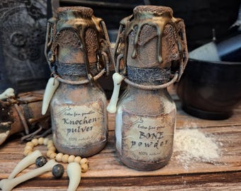 May include: Two small glass jars with cork stoppers and labels. The jars are filled with a white powder and have labels that read "Extra fein gemahlen Knochen pulver 100% natural" and "Extra fine grind Bone powder 100% natural".