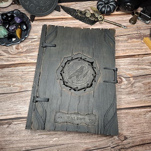 Könnte beinhalten: Ein dunkelgraues Holzbuch mit einem geschnitzten Rabenmuster und den Worten "Witch Book". Das Buch hat ein rustikales, gealtertes Aussehen mit dekorativen Schnitzereien und Metallverschlüssen, was auf ein Vintage- oder Fantasy-Thema hindeutet.