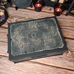 Könnte beinhalten: Eine dunkelgraue Holzkiste mit einem Used-Look. Der Deckel hat ein eingraviertes Design mit einem zentralen Halbmond- und Kronenmotiv, umgeben von keltischen Knotenmustern. Die Kiste ist rechteckig und scheint aus Holz zu sein.