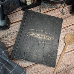 Könnte beinhalten: Ein dunkelgraues Kochbuch aus Holz mit dem Text "My Spellbound Recipes" auf dem Einband. Der Einband zeigt einen Hexenkessel und ein Hutdesign. Ein Holzlöffel liegt neben dem Buch auf einer Holzoberfläche.