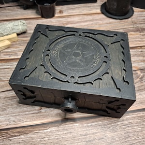 Puede incluir: Una caja de madera gris oscuro con un diseño de pentagrama en la tapa. La caja tiene cortes decorativos y un cierre de pestillo. La veta de la madera es visible y la caja parece hecha a mano. La estética general es gótica o mística.