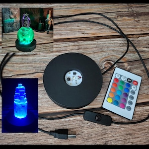 May include: A black LED light base with a remote control, USB cable, and a power switch. The remote has color options. The image also shows a glowing green orb and a blue crystal, both illuminated by the base.