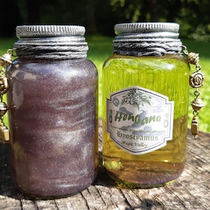 May include: Two glass bottles with silver lids and labels. The bottle on the left has a purple liquid with silver glitter. The bottle on the right has a yellow liquid and a label that reads "Henbane Hyoscyamus Magic Valley". Both bottles have a gold chain with charms attached.