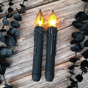 Könnte beinhalten: Zwei schwarze LED-Kerzen mit tropfenden Wachsdetails und flackernden bernsteinfarbenen Flammen. Die Kerzen stehen vor einem Holzgrund mit schwarzem Zierlaub und schaffen ein gotisches oder Halloween-Thema.