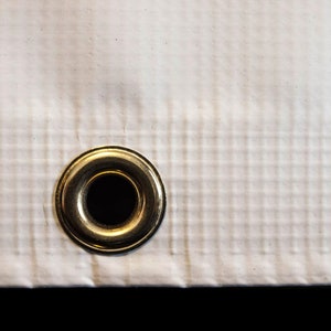May include: A close-up of a white fabric with a gold metal grommet. The fabric has a grid pattern.