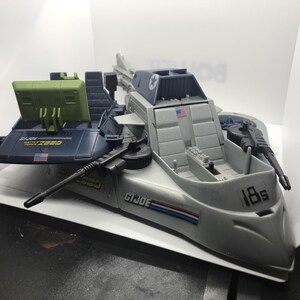 May include: A gray and dark blue G.I. Joe Battle Force 2000 toy vehicle featuring a light green radar screen, a small American flag decal, and the text 18s on the front. The plastic vehicle includes molded seating, black weapon attachments, and decals.