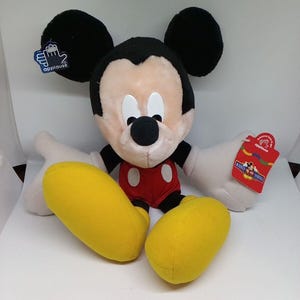 May include: A plush Mickey Mouse toy with black ears, a cream-colored face, and a black nose. He wears red shorts, yellow shoes, and white gloves. The toy has tags from the manufacturer, Applause.
