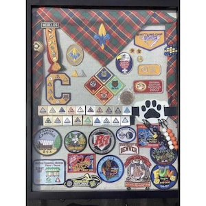 May include: A framed collection of Boy Scout memorabilia, including patches, pins, and a neckerchief. The display features badges with text like "WEBELOS," "BEAR," and "FRISCO ROUGHRIDERS." Various colors and shapes are visible, creating a nostalgic theme.