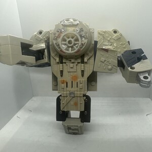 May include: A beige and gray plastic robot action figure with a circular cockpit-style chest piece, mechanical detailing, and articulated arms. The figure features a central wheel-like design on the torso and metallic silver accents on the hands.