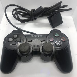 May include: A black Sony PlayStation DualShock controller featuring a directional pad, two analog sticks, four action buttons with triangle, circle, square, and cross symbols, plus Select, Start, and Analog buttons, connected by a black wired cable.