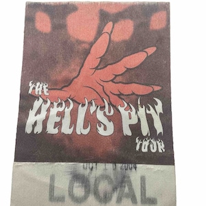 May include: A brown and red fabric patch with the text "The Hell's Pit Tour" and the word "LOCAL" printed on a white fabric tag below. The patch features a red hand with flames coming from the fingers.