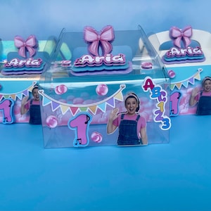 May include: Three decorated party favor boxes with the name "Aria" and the number "1". Each box has a pink bow, a picture of a woman, and bubble graphics. The boxes are on a blue surface.