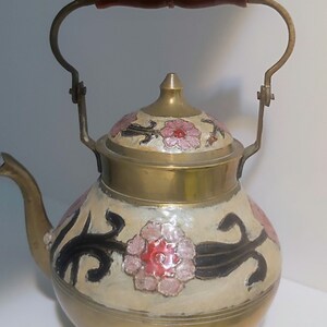 Könnte beinhalten: Eine Messing-Teekanne mit Holzgriff und floralem Design. Die Teekanne hat einen cremefarbenen Körper, der mit rosa Blumen und schwarzen Akzenten verziert ist. Der Griff ist dunkelbraun und Ausguss und Deckel sind aus Messing.