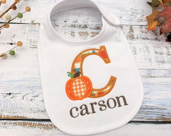 May include: White bib with a brown and orange patchwork letter C, a small orange pumpkin with a white and red checkered pattern, and the name "carson" embroidered in brown.