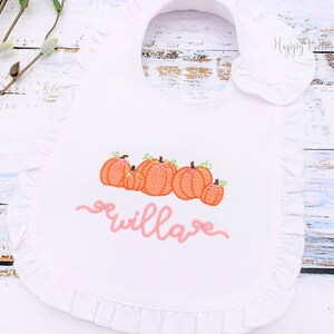 May include: White bib with a pink ruffle trim and embroidered pumpkins and the name "willa" in pink.