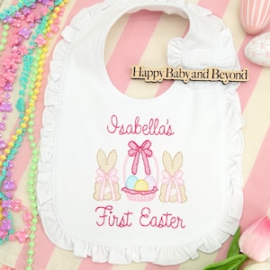May include: White baby bib with a ruffled edge, embroidered with the text "Isabella's First Easter". The bib features a basket with Easter eggs and two bunnies with bows. The bib is on a pink and white striped background.