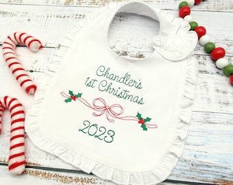 May include: White baby bib with a ruffled edge, embroidered with "Chandler's 1st Christmas 2023" in green script. A red ribbon and holly design is embroidered below the text. Two red and white candy canes are on the left.