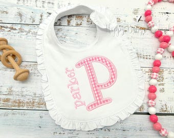 May include: White baby bib with a pink gingham letter "P" and the name "Parker" embroidered in pink. The bib has a ruffled edge. A wooden rattle and a pink and white bead garland are also in the image.