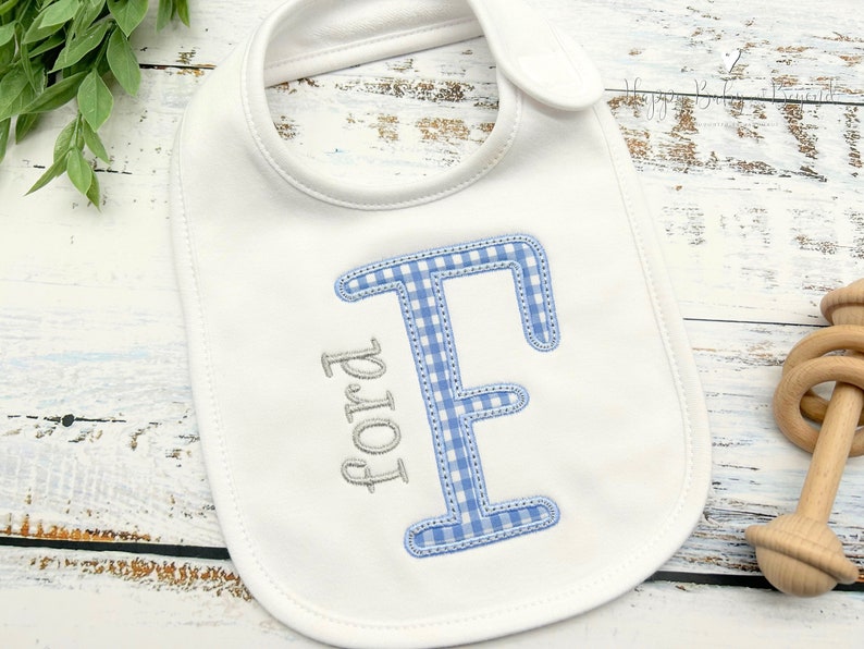 May include: White baby bib with a blue and white gingham letter E and the name Ford embroidered in gray.
