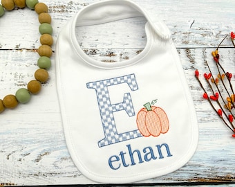 May include: A white baby bib with a blue and white checkered letter E, a small orange pumpkin, and the name "ethan" embroidered in blue thread.
