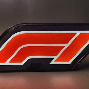 May include: A black and orange Formula 1 logo, featuring the iconic "F1" emblem. The logo has a black border with a white outline around the orange design. The image is a close-up shot, highlighting the logo's shape and color.