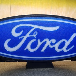 May include: A blue oval sign featuring the white 'Ford' logo. The sign has a black border and is mounted on a black stand. The background is a light yellow colour.